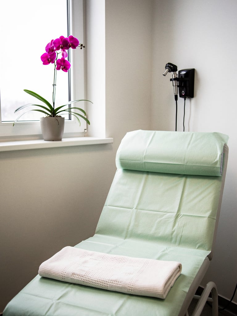 Canto do consultório com maca coberta de papel verde-claro, otoscópio na parede e orquídea magenta no peitoril